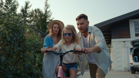 Happy family on vacation spending carefree time together outside. Attractive dad and pretty mom with daughter having fun and walking in nature. Leisure of excited girl riding bicycle in cottage garden