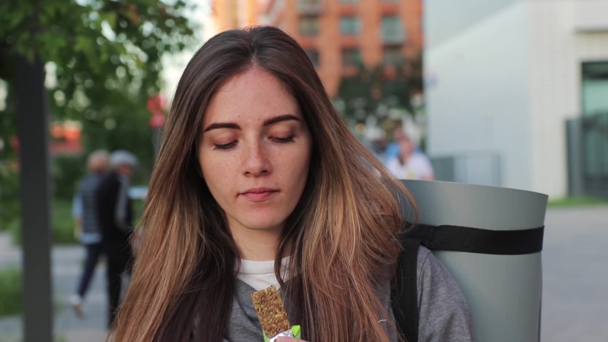 woman with long hair eats a granola bar after training with a sports mat on the back in the busy street Royalty-Free Stock Footage #1060089797