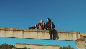 Scary female vampire and grim reaper with scythe in black cloak talking, relaxing and getting ready for evil deeds while sitting on concrete beam on halloween over blue sky background. - Powered by Shutterstock - Get 15% off with code: PIKWIZARD15