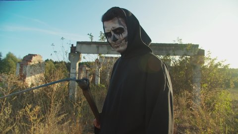Portrait of creepy grim reaper with rustic scythe standing in ruins of abandoned building in rays of glowing setting sun, looking with mysterious cold eyestare while haunting lost soul on halloween.