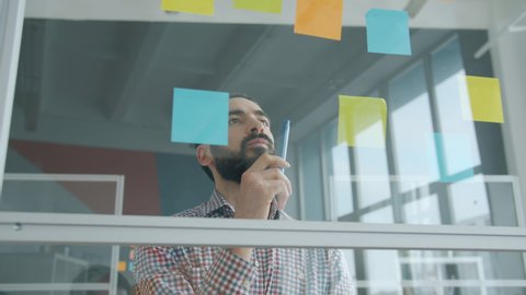 Slow motion of young mixed race businessman working with sticky notes on glass wall in office focused on creative work. Businesspeople and job concept.