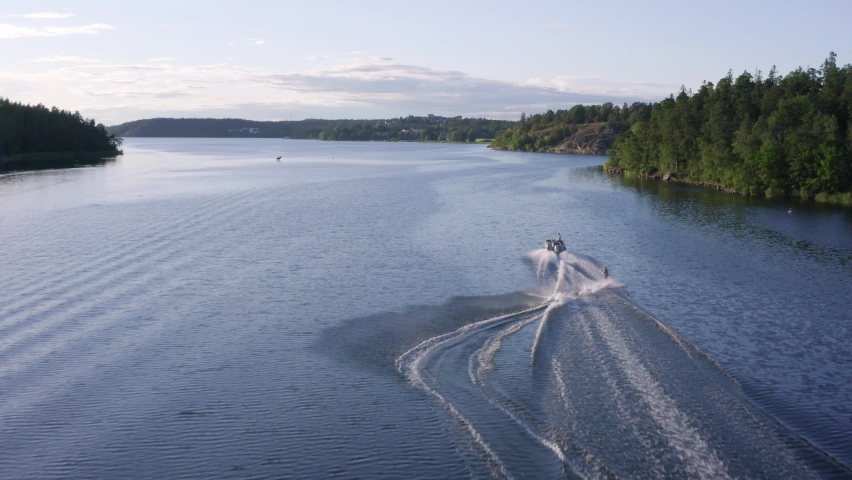 Water ski on lake in aerial drone shot. View of blue water and forest. Some houses at ocean front. Strong female boarder riding waves behind motor boat