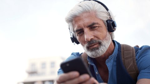 Portrait of mature man with headphones sitting outdoors in city, using smartphone.