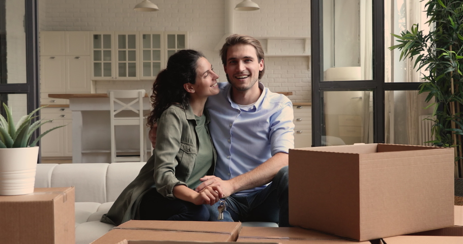 Happy young couple sit on sofa near of heap belongings in cardboard boxes, wife showing to camera bunch of keys, symbol of new modern home. Excited homeowners portrait, real estate and tenancy concept Royalty-Free Stock Footage #1060104272