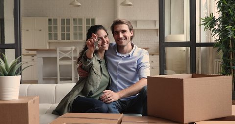 Happy young couple sit on sofa near of heap belongings in cardboard boxes, wife showing to camera bunch of keys, symbol of new modern home. Excited homeowners portrait, real estate and tenancy concept