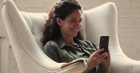 Woman resting in cozy soft armchair with smartphone. Pretty smiling female looks at device screen use selfie cam virtual makeover modern application on cell, photo-sharing app. Social media fun