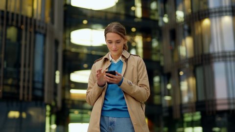 Zoom in panning shot of beautiful young woman in trench coat standing in city street, text messaging on cell phone and looking away dreamily