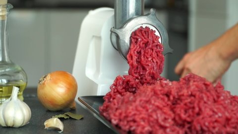 Chef hands put meat in electric meat grinder on background with onion, garlic and olive oil. Preparation of minced fresh beef for burgers. Food background.
