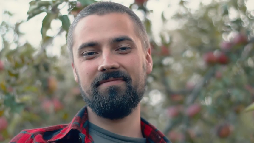 Portrait of a smiling bearded caucasian gardener against the background of trees with apples. Organic farm concept. Close-up, slow motion