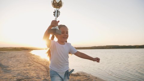 Happy kid winner run along beach at sunset with cup in his hand above his head. Kid with winner cup. Success of the victory. Kid with cup runs to victory. Winner concept. Happy kid winning with cup