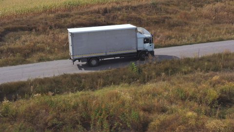 Aerial shot of truck with cargo trailer driving on road and transporting goods. Flying over delivery lorry moving along highway passing near fields in countryside. Scenic nature scene. Side view
