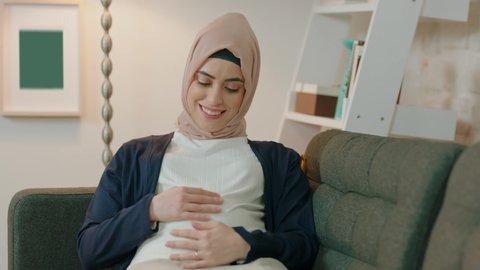 Pregnant woman feels happy at home while loving her child. Young muslim expectant mother holding her pregnant belly. Maternity prenatal care and female pregnancy concept.