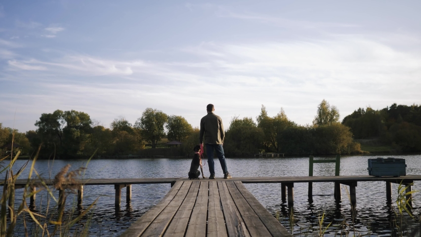 An amazing shot of a young man with his pet dog standing on the edge of the pier and admiring the beauty of the lake at sunset.