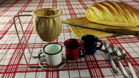 Coffee being filtered through a cloth strainer in enameled cups and homemade bread in the background on a checkered tablecloth. Making coffee with an cloth strainer also known as Mariquinha in Brazil