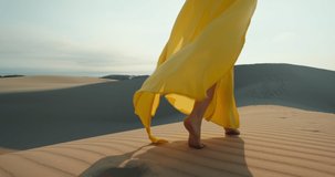 Beautiful fashion video. Bright dress is flying by the wind in slow motion with blue sky on background. Commercial footage for clothing business. Cinematic modern shot of woman walking by desert sand
