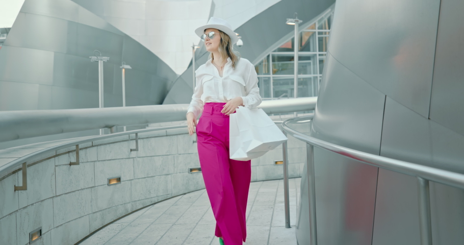 Slow motion fashion model. Pretty smiling blonde confidently walks by the street with white shopping bags, touches trendy white hat. Joy of life, lifestyle, fashion blogger, catwalk. 4K slow motion