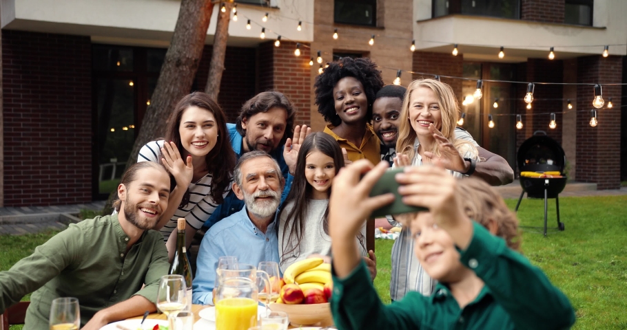Mixed-races happy family at party dinner outdoor in yard smiling and posing to smartphone camera while small boy taking selfie photo. Multi ethnic people making photos together at barbeque Celebration Royalty-Free Stock Footage #1060139060