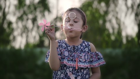 A cute and adorable little girl child is blowing soap water bubbles in the park or garden. 