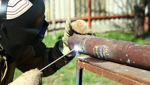 male welder in protective suit and mask is welding a thick steel pipe with an electrode on an iron table with an electrode outdoors, close up, the concept of repair work