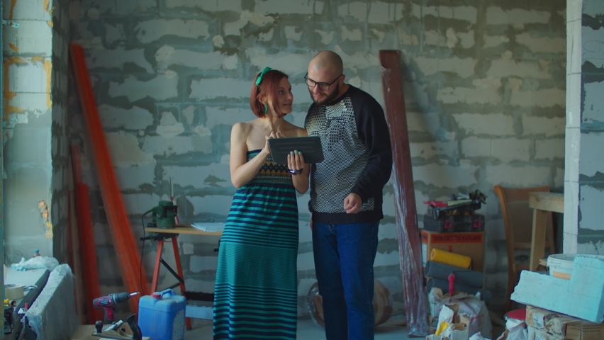 Young excited couple planning their new house renovation looking at blueprints on tablet computer standing at construction site. Happy millennial family hugging. 
