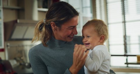 Authentic shot of an young happy smiling mother is keeping in arms her toddler baby boy and having fun to dance together in a kitchen at home.