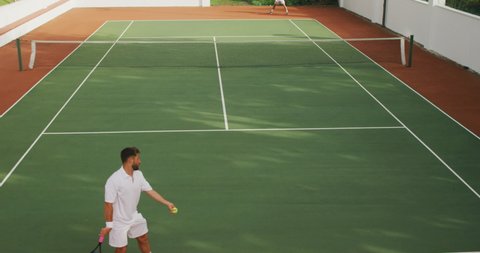 A Caucasian and a mixed race men wearing tennis whites spending time on a court together, playing tennis on a sunny day, holding tennis rackets, one of them hittig a ball, in slow motion.