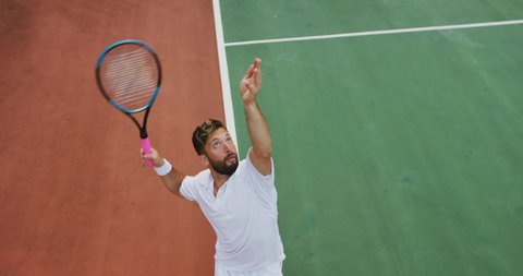 A mixed race man wearing tennis whites spending time on a court, playing tennis on a sunny day, holding tennis racket, hittig a tennis ball, in slow motion.