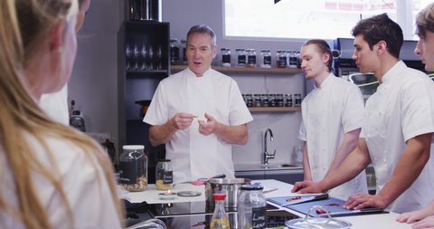 Professional Caucasian male chef wearing chefs whites in a restaurant kitchen teaching a group of male trainee chefs, showing them how to cook in slow motion