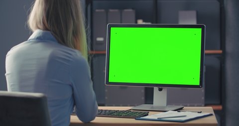 Back view of female office worker in blue shirt sitting at desk, looking at green computer screen and taking notes. Space for text and advertisement. Concept of technology and business.