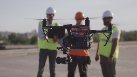 Modern drone flying in air near male contractors with remote control and laptop on bridge construction site