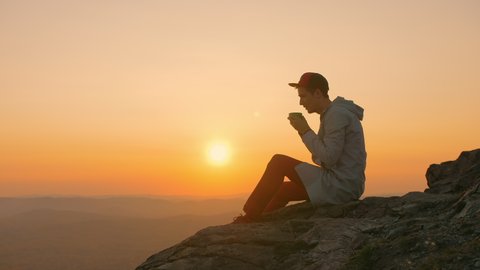 Man tourist sits on the mountain top. drinks tea from a mug. enjoying the sunset and picturesque landscape. Hipster hiker sitting on edge of cliff, relaxed on stone, looks into distance and dreaming.