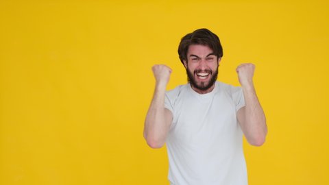 Handsome bearded young guy 20s in denim shirt white t-shirt isolated over yellow background in studio. People sincere emotions, lifestyle concept. Looking at the camera doing winner gesture, say yeah.
