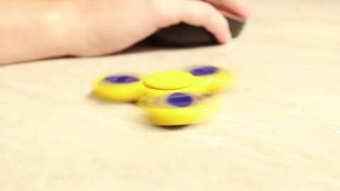A yellow fidget spinner is spinning on a computer table, with a man working at a computer in the background. Close up.
