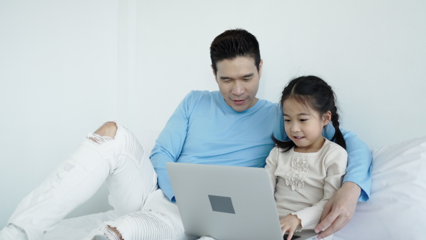 Happy Asian father and daughter are playing with a laptop in house. Family activities teach children the use of technology in education. Online shopping. Meeting video conference with people