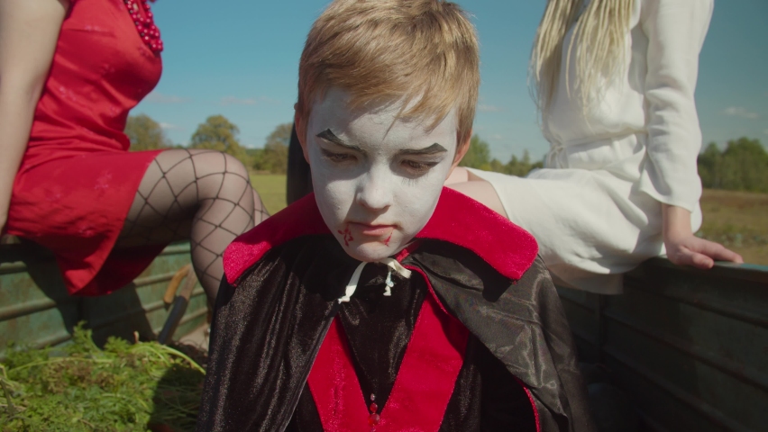 Upset preadolescent boy with halloween make-up in vampire costume, sitting in mini tractor trailer with other females souls while grim reaper in shroud transporting souls of deceased to afterlife.