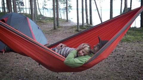 Active family outdoors, travel and adventure with child. Father and son hikers travellers sitting in tourist hammock near camping tent in the forest.: film stockowy