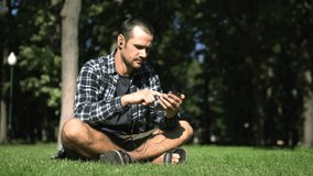 Young handsome guy in a plaid shirt leafs through the feed of social networks while sitting on the grass in the park. A young guy reads the feed of social networks on his smartphone while sitting on
