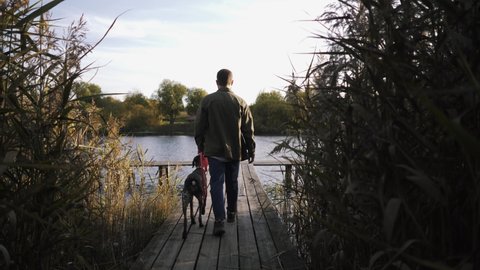 Side view of a young guy sitting with his pet dog on the pier and admiring the beauty of the autumn lake. A man looks kindly into the eyes of his dog.