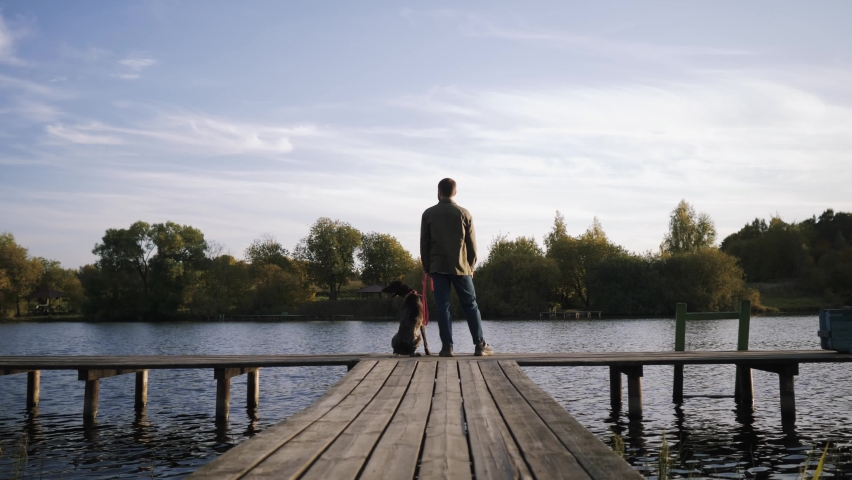 An amazing shot of a young man with his pet dog standing on the edge of the pier and admiring the beauty of the lake at sunset.