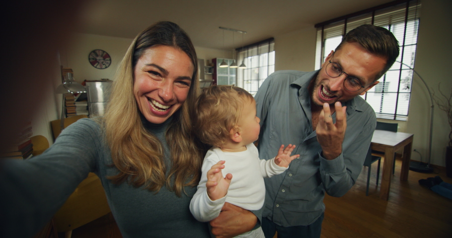 A happy smiling family with toddler baby boy is making a selfie or video call to friends or relatives while having a breakfast together in a living room at home. Royalty-Free Stock Footage #1060179302