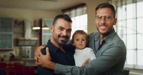 Authentic shot of young happy homosexual male gay family with adopted son toddler baby boy is smiling in camera in a kitchen at home.
