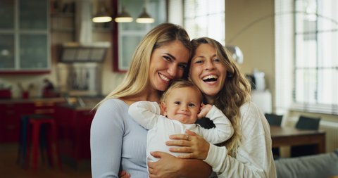 Authentic shot of young happy homosexual female gay family with son toddler baby boy is smiling in camera in a kitchen at home.