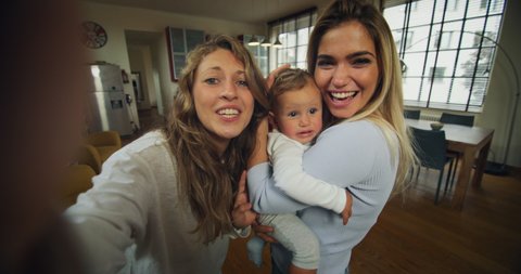 Authentic shot of young happy homosexual female gay family with son toddler baby boy is making a selfie or video call to friends or relatives in a living room at home.