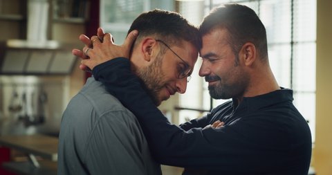 Authentic shot of young happy smiling married homosexual male gay couple is enjoying time together is hugging and kissing as sign of timeless love in a kitchen at home. 库存视频