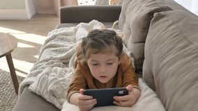 A little girl with a beautiful hairstyle attentively watches cartoons online on her mobile phone lying on the sofa and covered with a blanket
