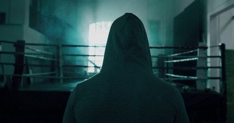 Cinematic slow motion back view shot of a professional young man wearing grey hoodie is entering in a boxing ring for starting his workout.