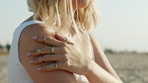 Young unrecognizable woman caressing her shoulder, strokes body on sandy beach. Beautiful girl touching her forearm. Hand with fashionable rings. Jewelry, fashion, healthy skin concept