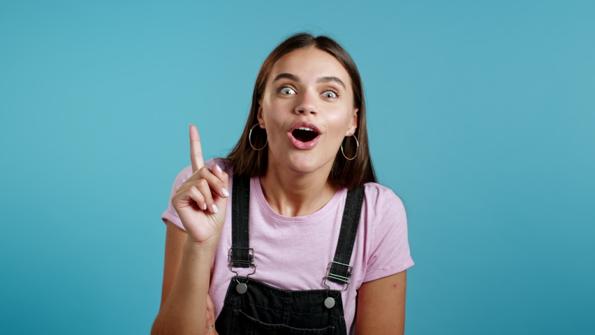 Smiling happy girl showing eureka gesture. Portrait of young thinking pondering woman having idea moment pointing finger up on blue studio background.  | Shutterstock HD Video #1060185275