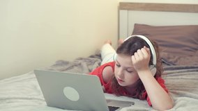 A beautiful girl lying on the bed is studying online with white headphones. School online during a pandemic.