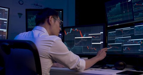Asian business man is sitting at desk with monitors showing line graphs, charts. Alone male is analyzing stock market trading, strategic planning project of marketing data at late night. Dolly shot.